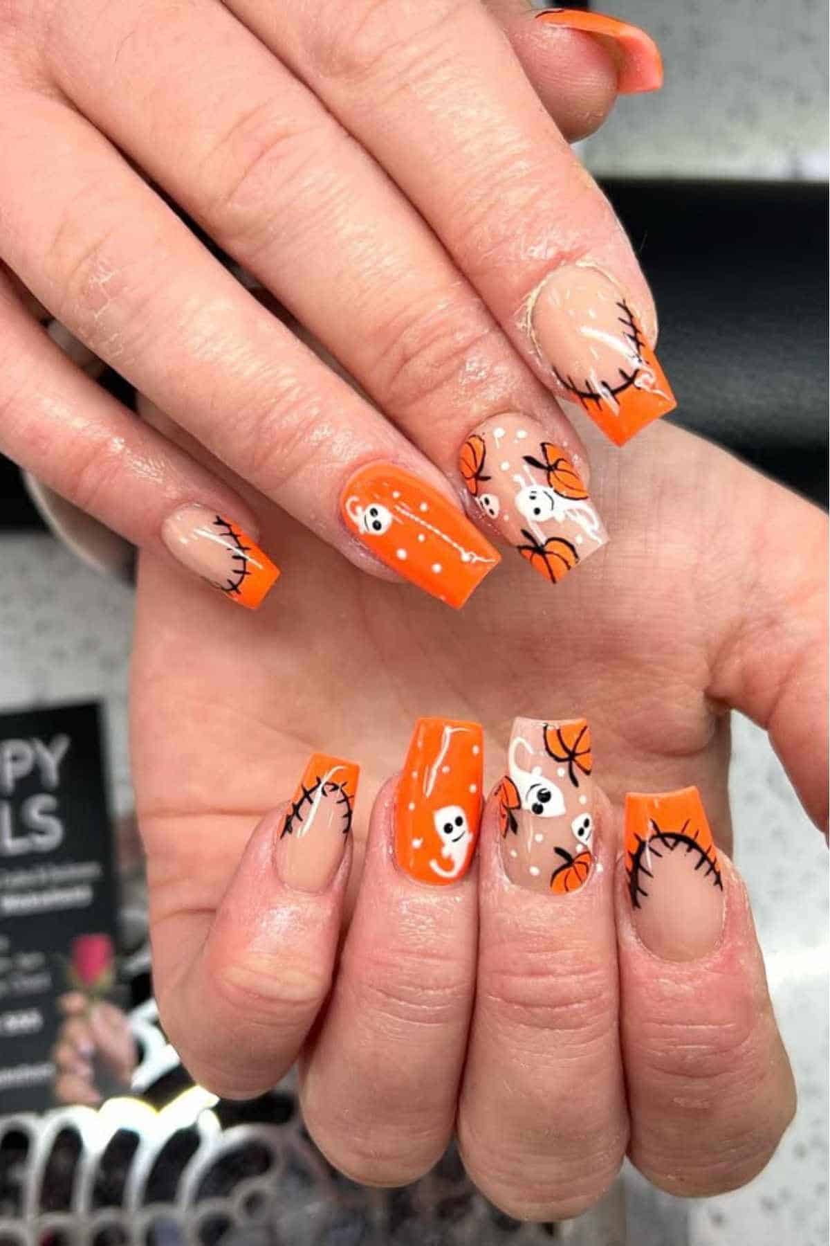 Hands with long square nails featuring orange tips and Halloween-themed nail art, including ghosts, pumpkins, and black stitched details.