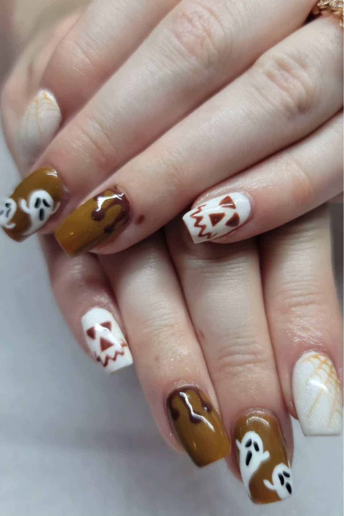 Close-up of hands with brown and white Halloween-themed nail art featuring ghosts, jack-o'-lantern faces, and drips.