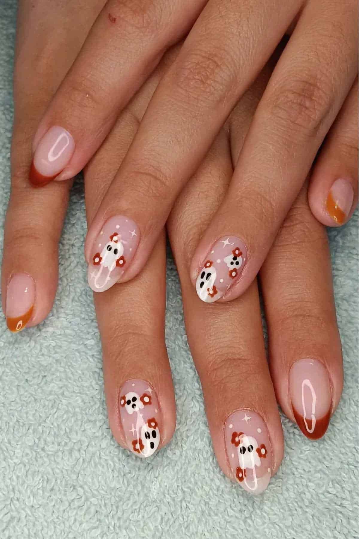 Hands with short nails featuring a clear base, orange French tips, and white ghost designs with black eyes and mouths, along with small orange and white flowers.