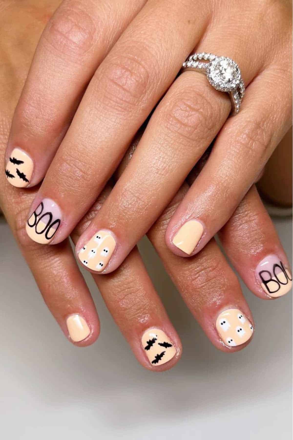 Hands with pale peach Halloween-themed nail art featuring the word "BOO," black bats, and small white ghosts with dotted eyes; a diamond ring is worn on one finger.