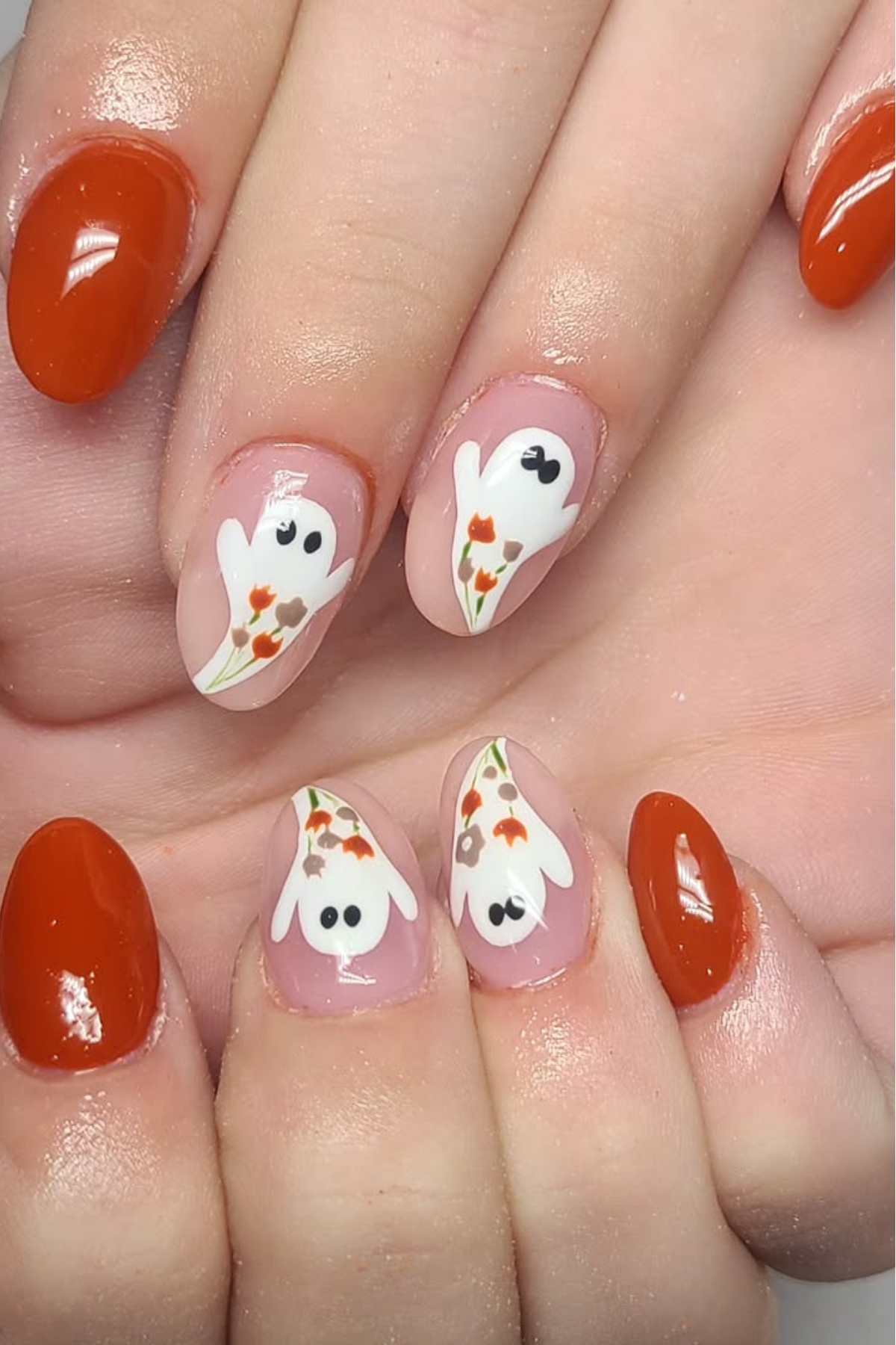 Fingernails painted with a mix of solid burnt orange polish and designs of white ghost shapes with black eyes and floral accents on a nude base.