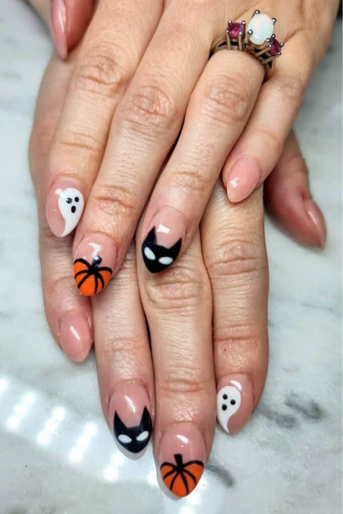 Hands with Halloween-themed nail art featuring black cats, orange pumpkins, and white ghosts on a marble surface.
