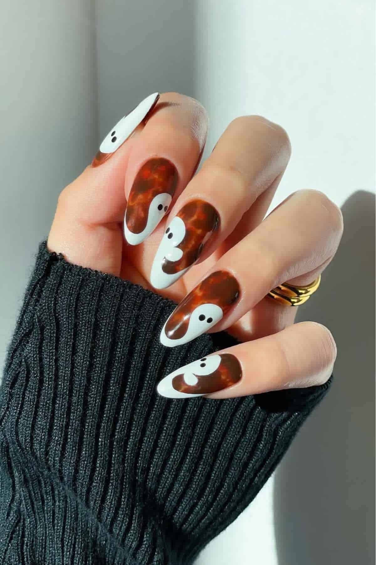 A hand with long almond-shaped nails featuring a brown tortoiseshell pattern and white abstract faces, partially covered by a black sweater sleeve.