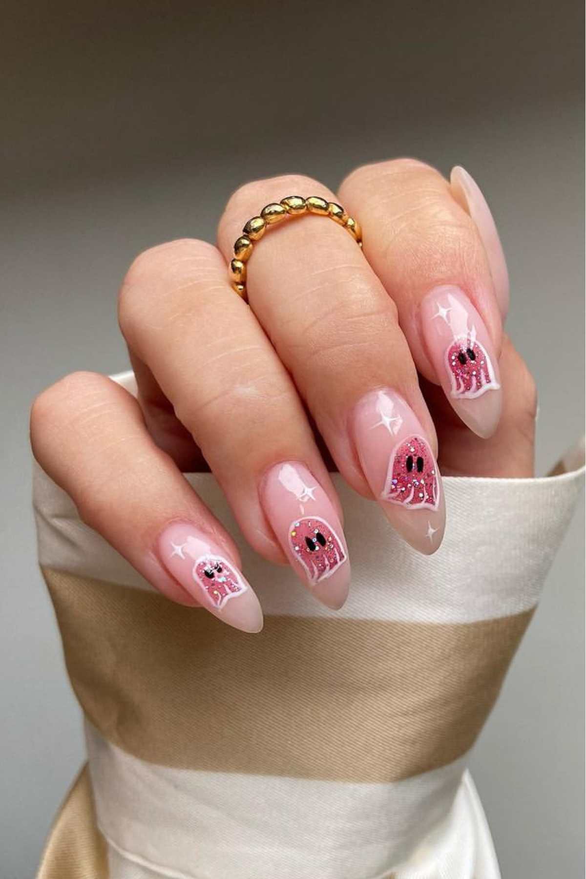 A hand with almond-shaped nails featuring a pink ghost design and sparkles on a light nude base; the person wears a gold beaded ring.