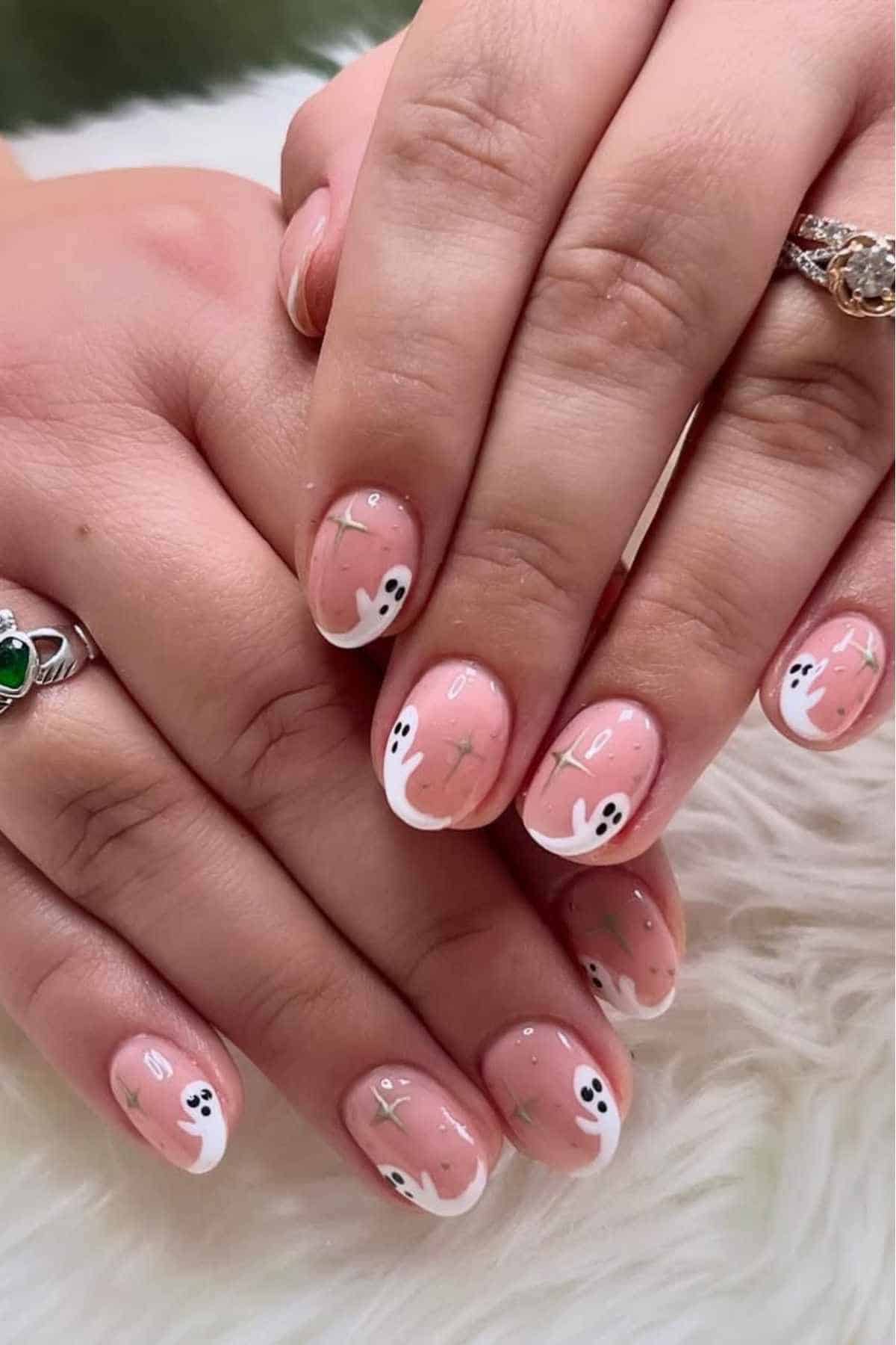 Hands with short, almond-shaped nails painted with nude polish and white ghost designs, featuring minimalist details and accent rings on two fingers.