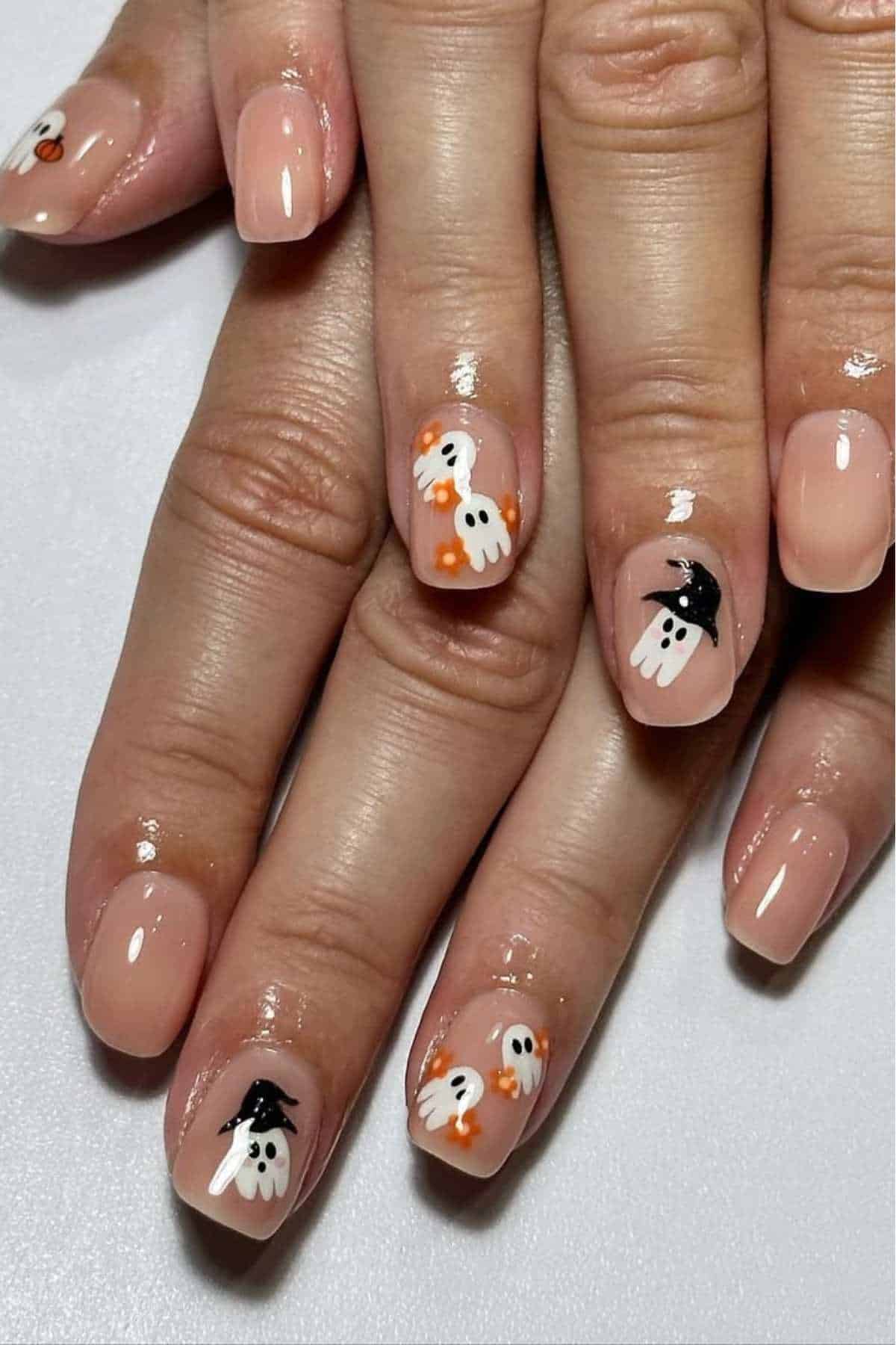A close-up of hands with nude-colored nails featuring painted white ghost designs, some with orange accents and black hats, on a few nails.