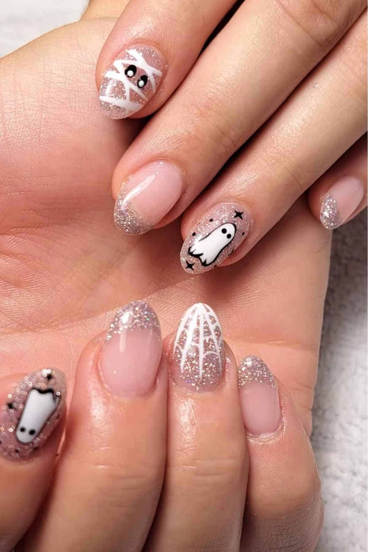 A close-up of hands with manicured nails featuring glitter tips, white spider web designs, and ghost and skull nail art.