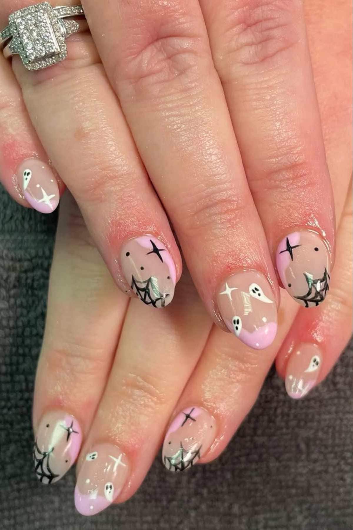 Hands with pink-tipped nails featuring Halloween-themed nail art, including black webs, small white ghosts, and stars. A diamond ring is worn on one finger.