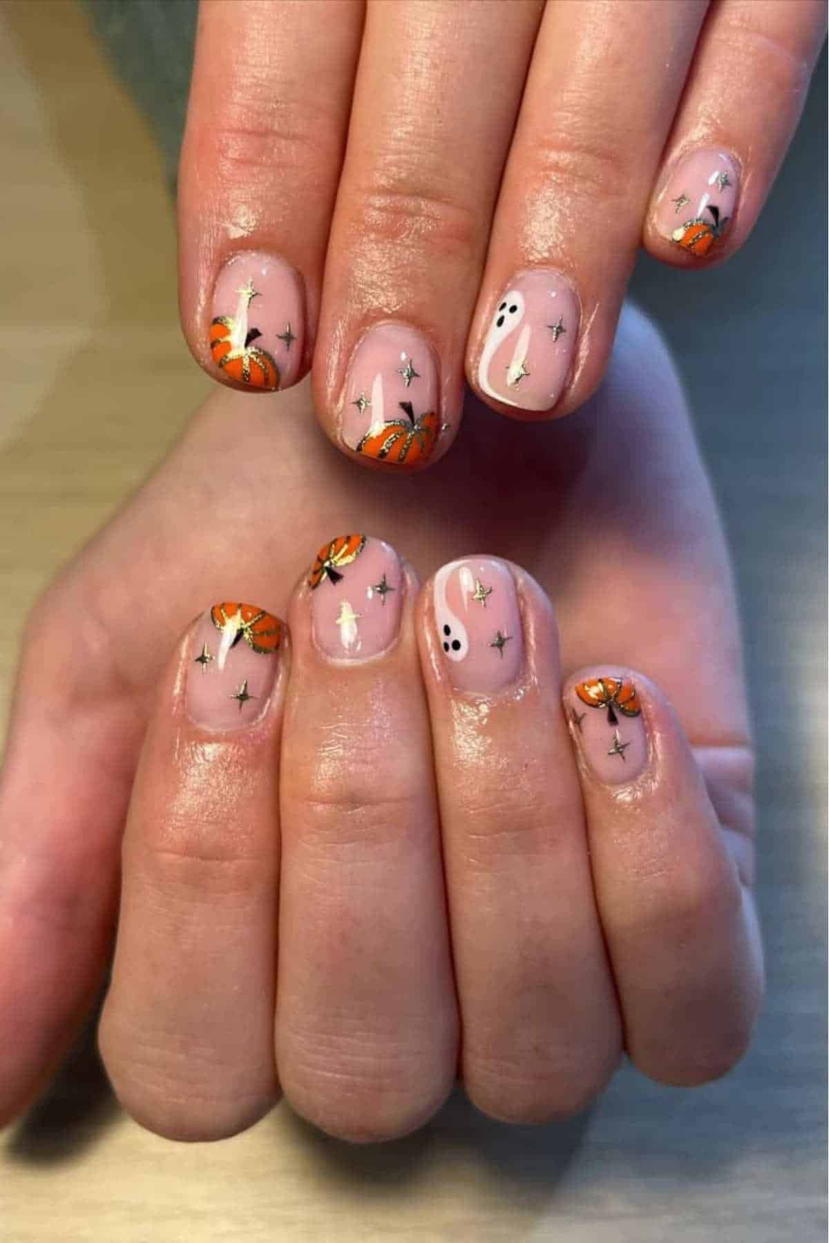 Hands with short, natural nails featuring Halloween-themed nail art, including pumpkins, ghosts, and black stars on a nude base.