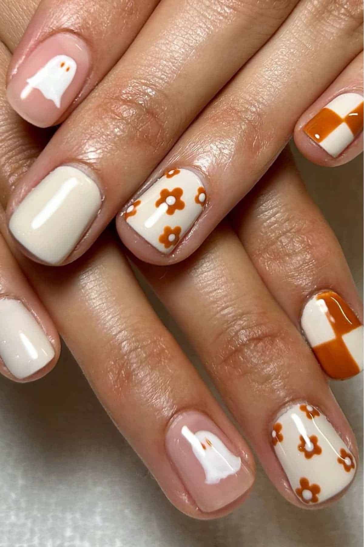 Close-up of hands with short, almond-shaped nails painted in a cream base, featuring orange and white designs: ghosts, flowers, and checkerboard patterns.