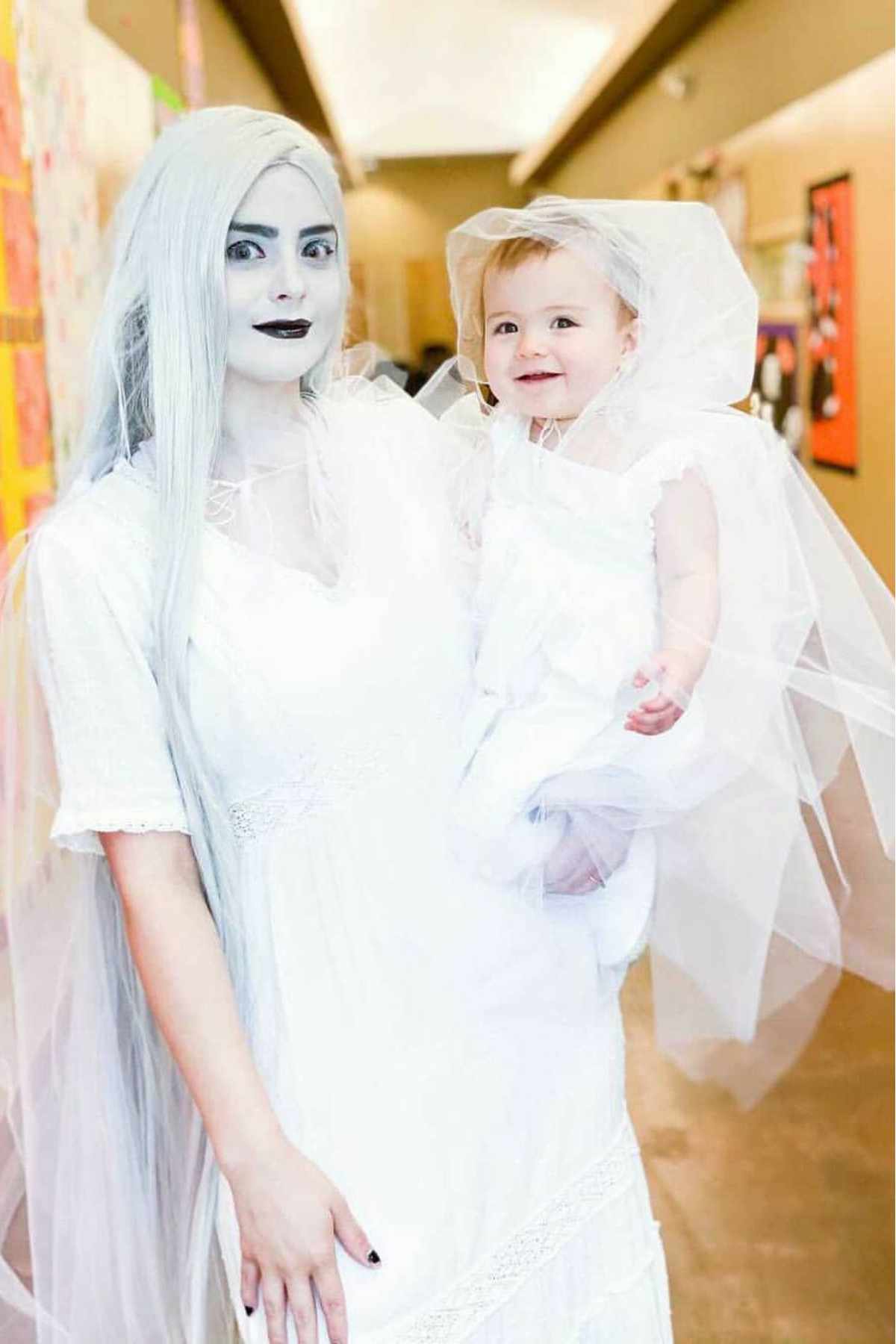An adult and a child dressed in white costumes, with the adult wearing white makeup and a long white wig, standing indoors in a hallway.