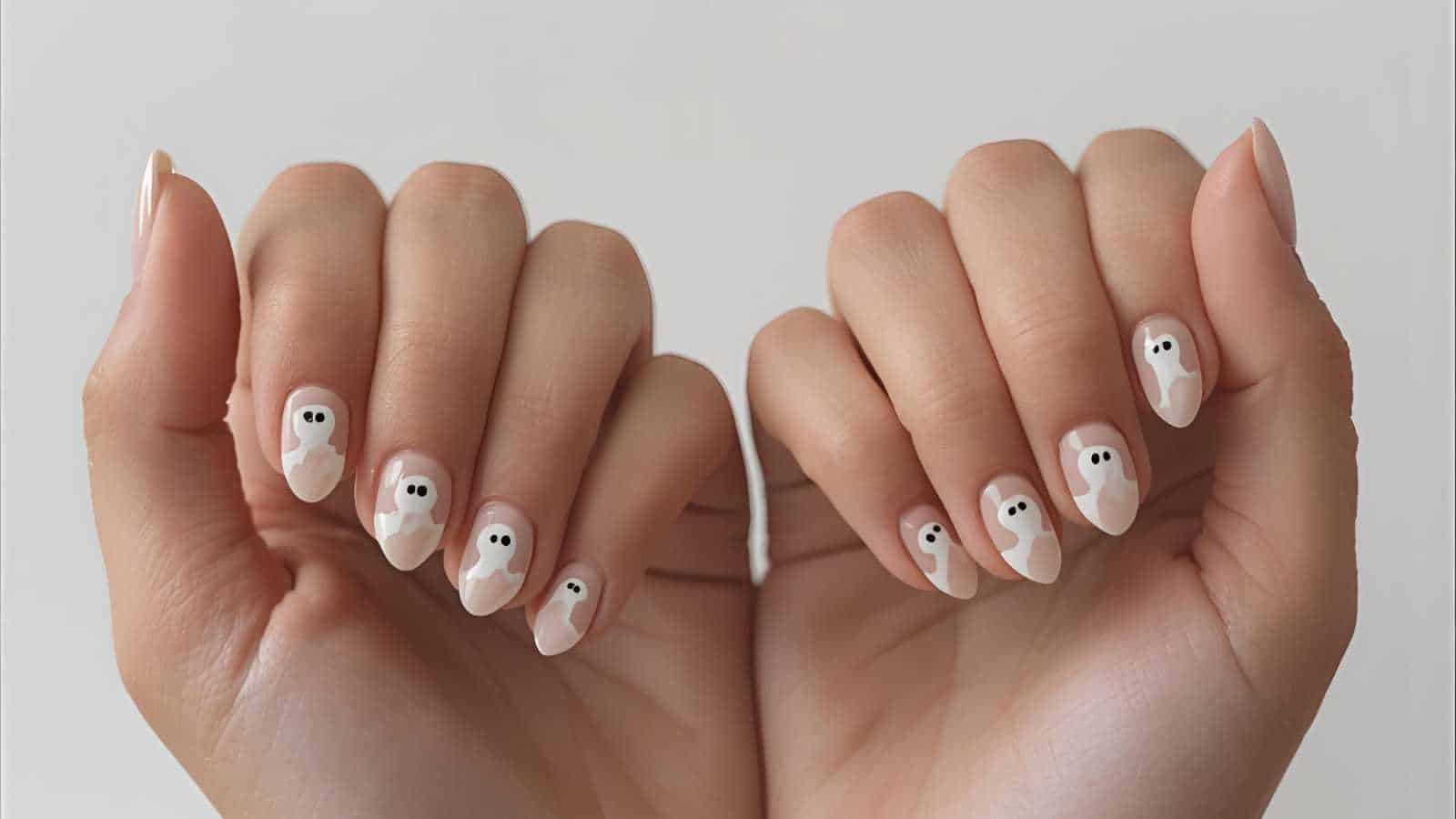 A pair of hands with nude-colored nails featuring white ghost designs with black eyes on each nail.
