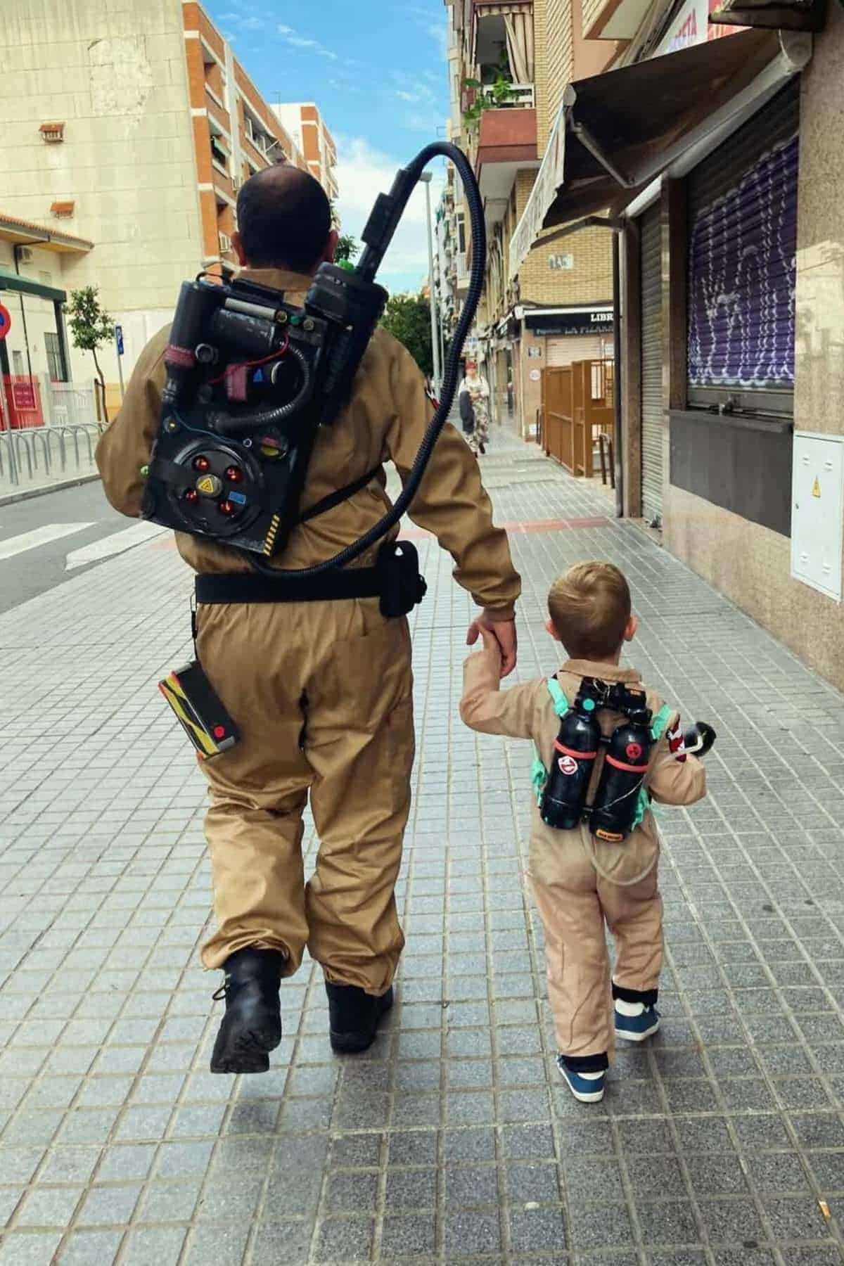 An adult and a child wearing matching Ghostbusters costumes and proton pack props walk hand in hand down a city sidewalk.