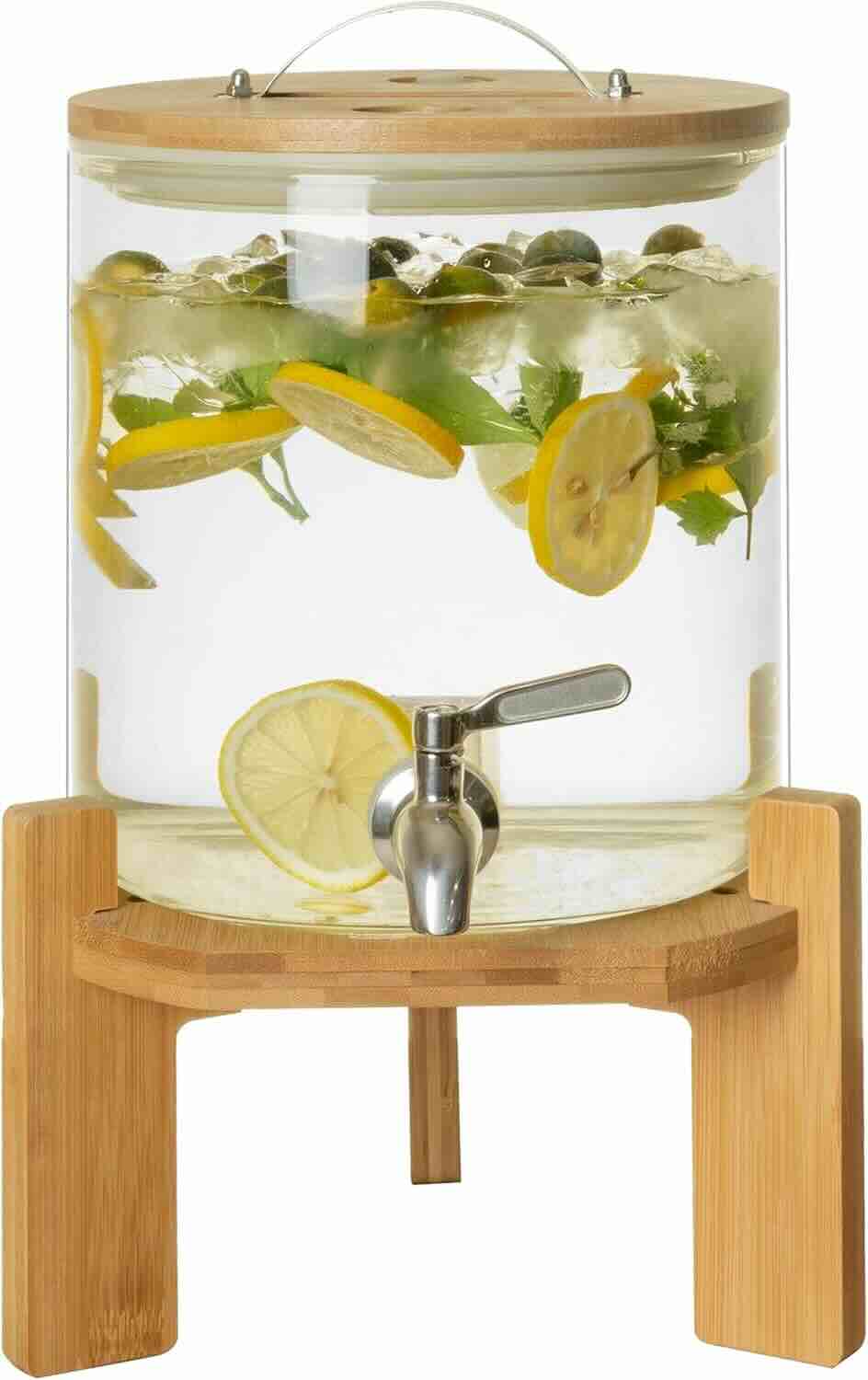 A glass drink dispenser with a wooden lid and stand, filled with water, lemon slices, herbs, blueberries, and ice, featuring a metal spigot for pouring.