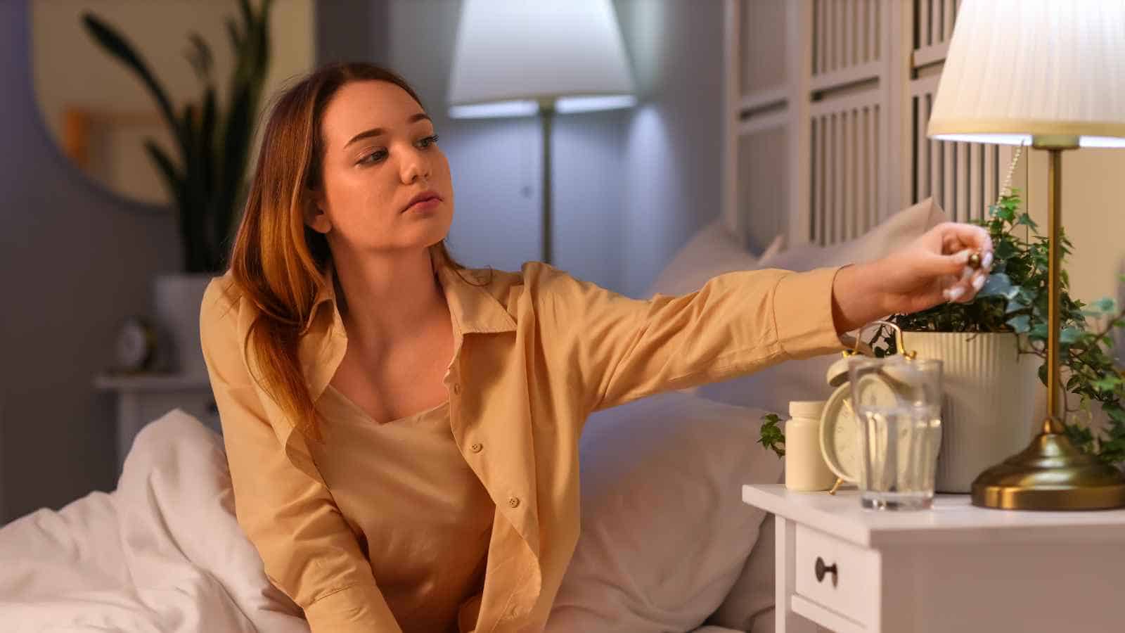 A woman sitting in bed reaches to turn off a bedside lamp; a glass of water, plant, and alarm clock sit on the nightstand.