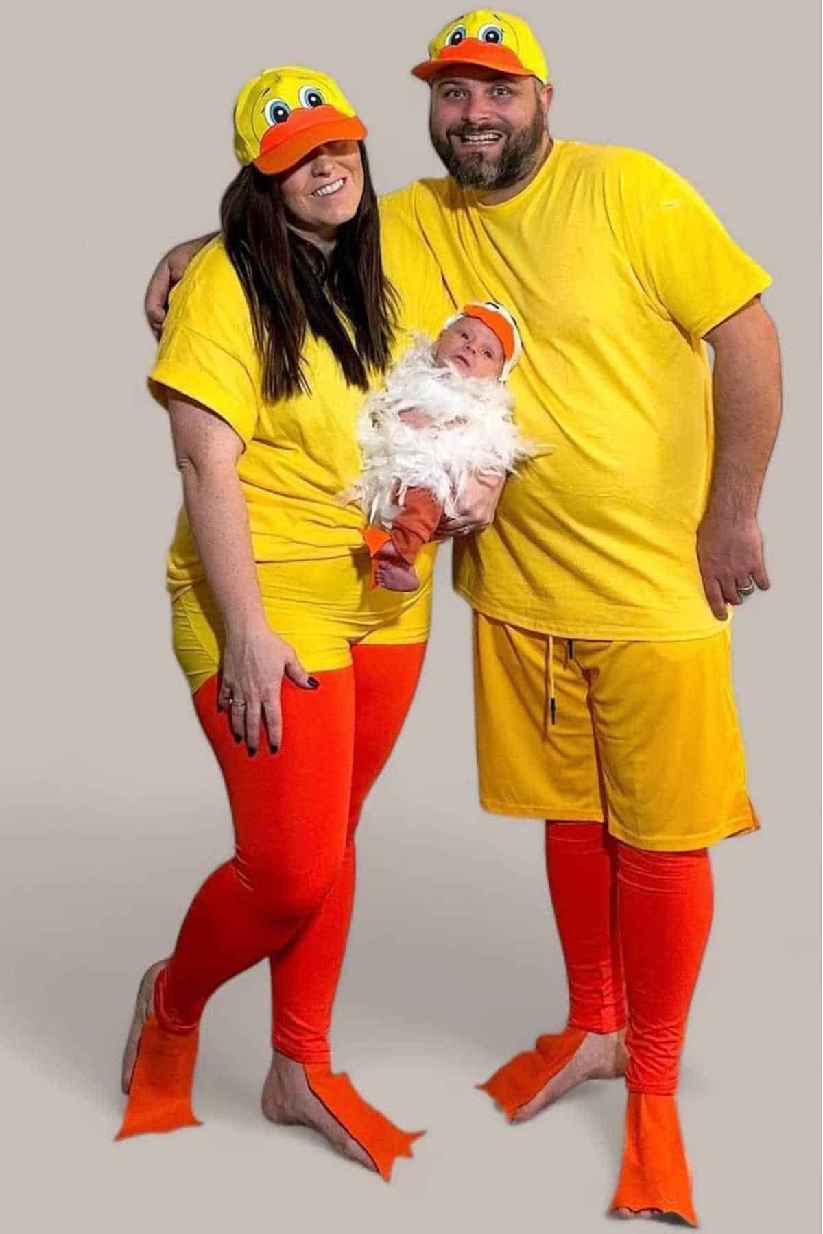 Two adults and a baby are dressed in matching yellow duck-themed costumes, including yellow shirts, hats with duck faces, orange leggings, and orange duck feet.