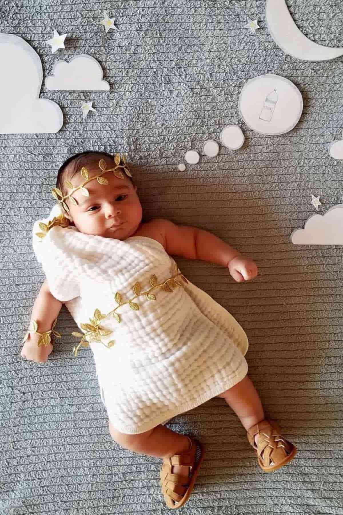 A baby dressed in a white cloth and sandals lies on a gray rug with paper clouds, moon, and thought bubbles, appearing to dream of a baby bottle.