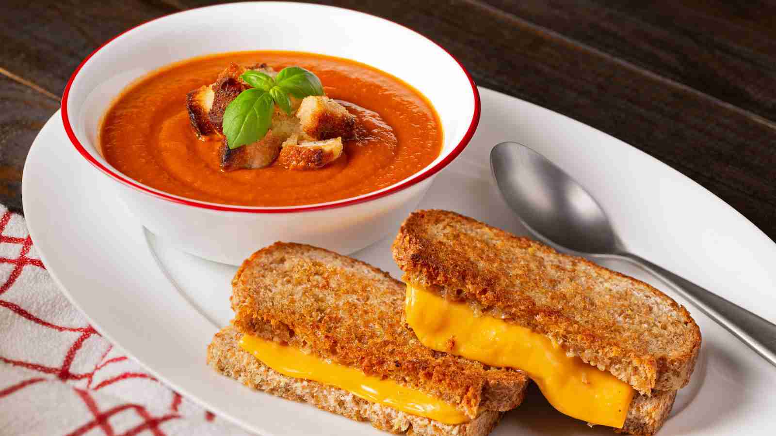A bowl of tomato soup topped with croutons and basil is served on a plate with a grilled cheese sandwich and a spoon.