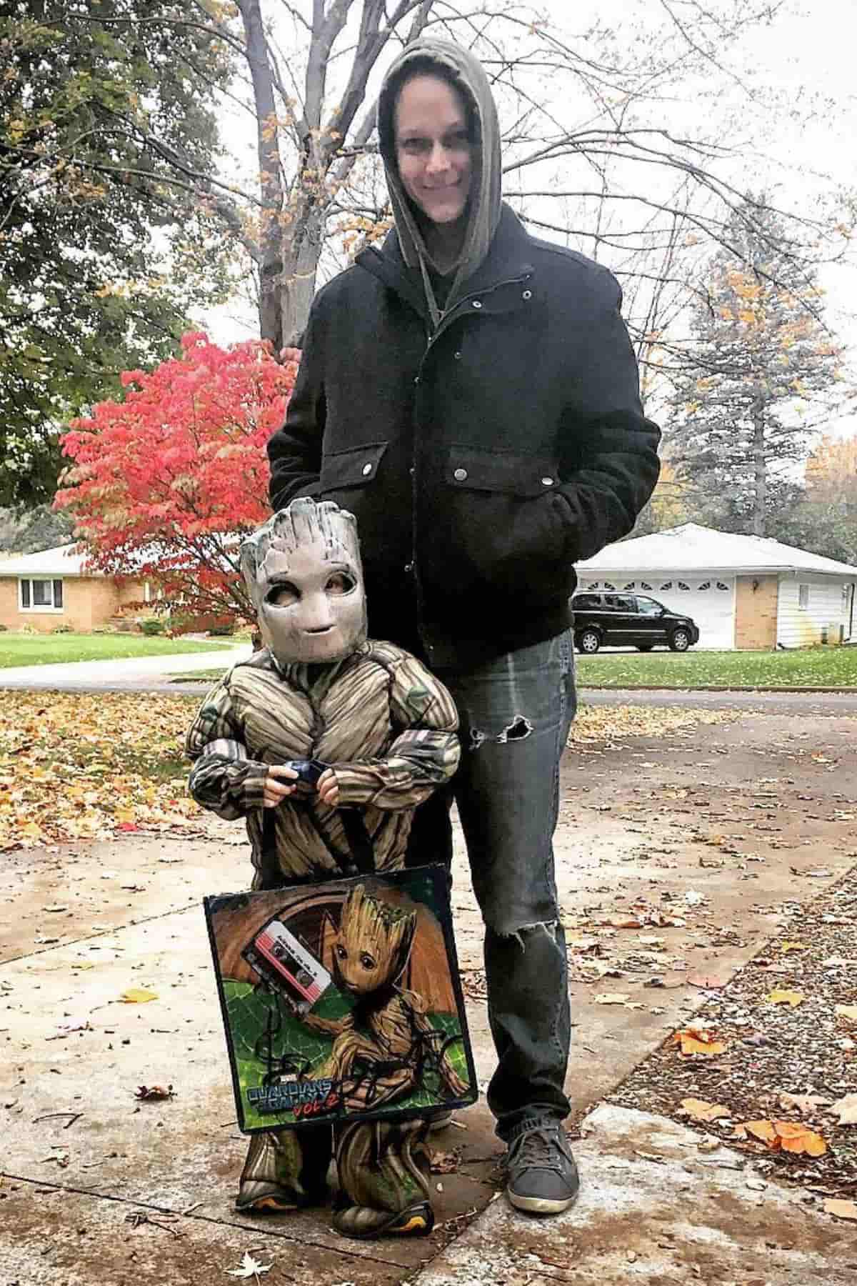 An adult stands behind a child dressed in a Groot costume holding a Groot-themed bag, outdoors in a suburban neighborhood during fall.