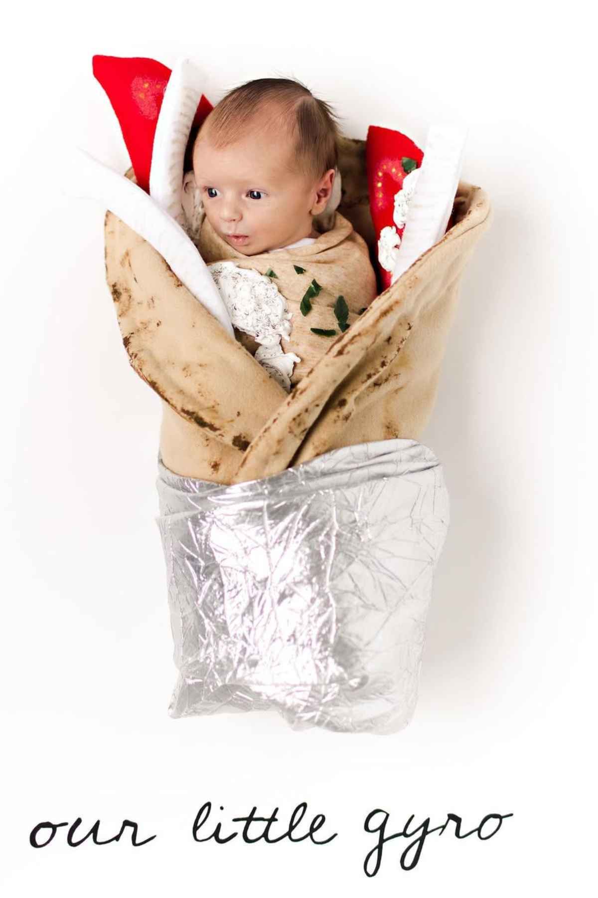 A baby is wrapped in a costume resembling a gyro sandwich, with felt toppings and a metallic wrap, on a white background. Text below reads "our little gyro."