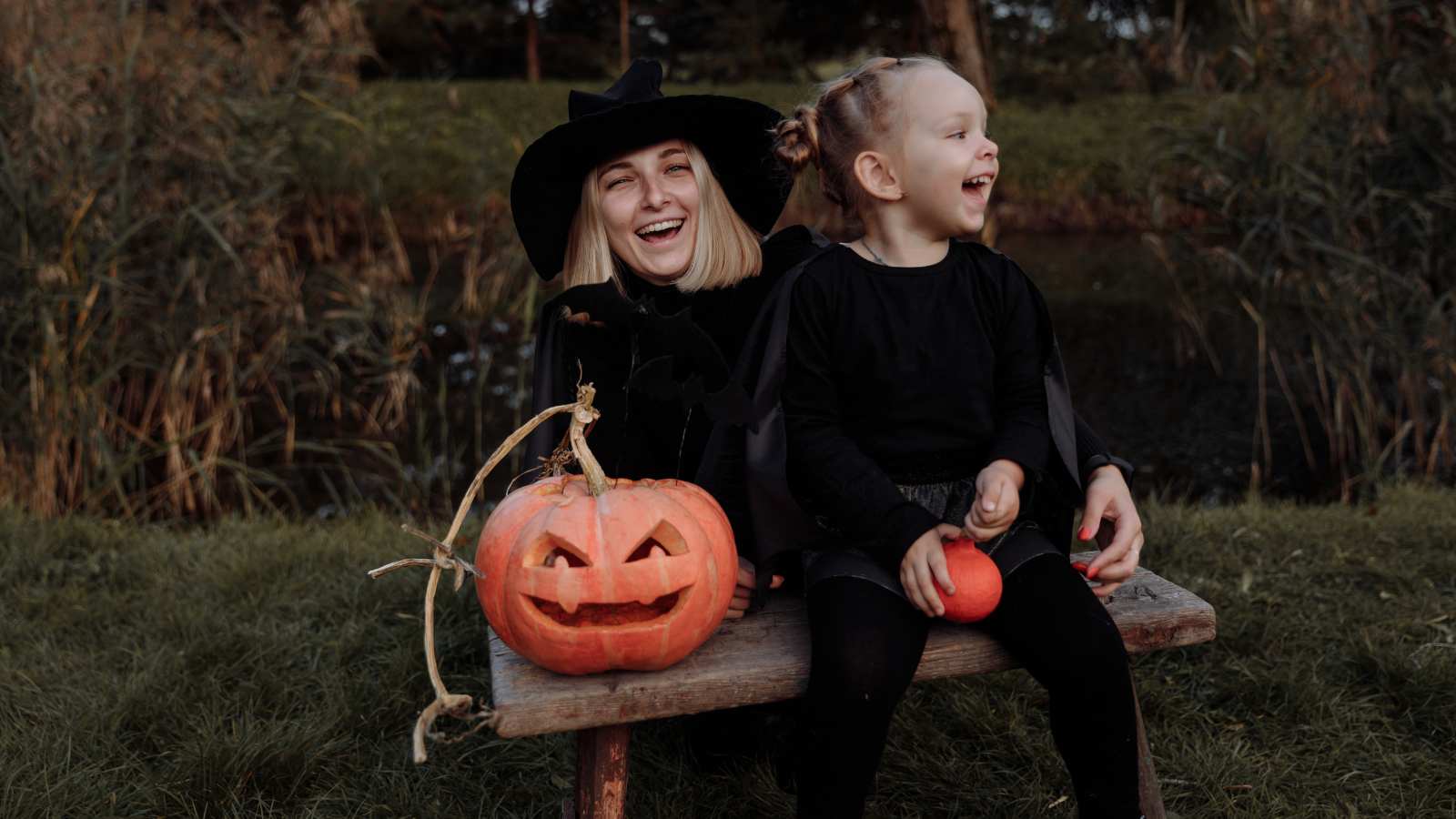 A mom and daughter in black costumes sit on a bench outdoors with carved and uncarved pumpkins, smiling and laughing.