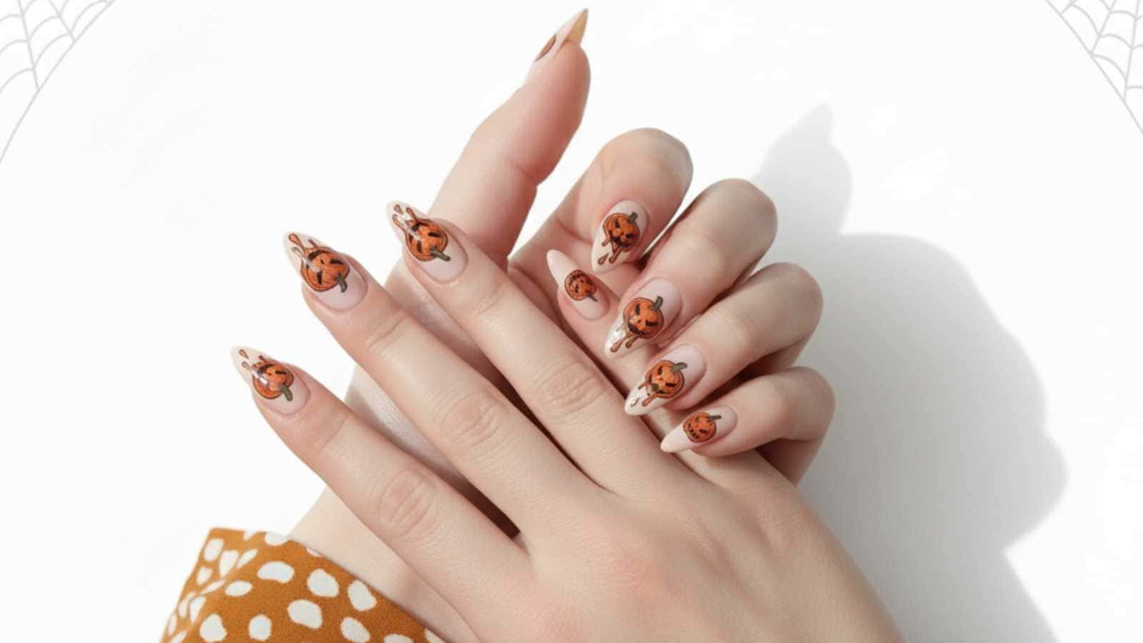 Hands with long fingernails painted with nude polish and decorated with halloween pumpkin nail designs, featuring orange jack-o'-lanterns, resting on a white surface.