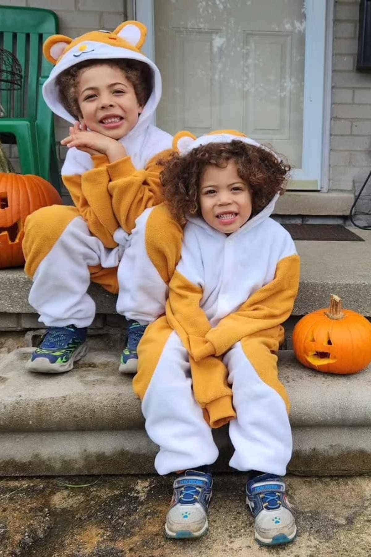 Two children in orange and white animal costumes sit on steps with carved pumpkins beside them in front of a house.