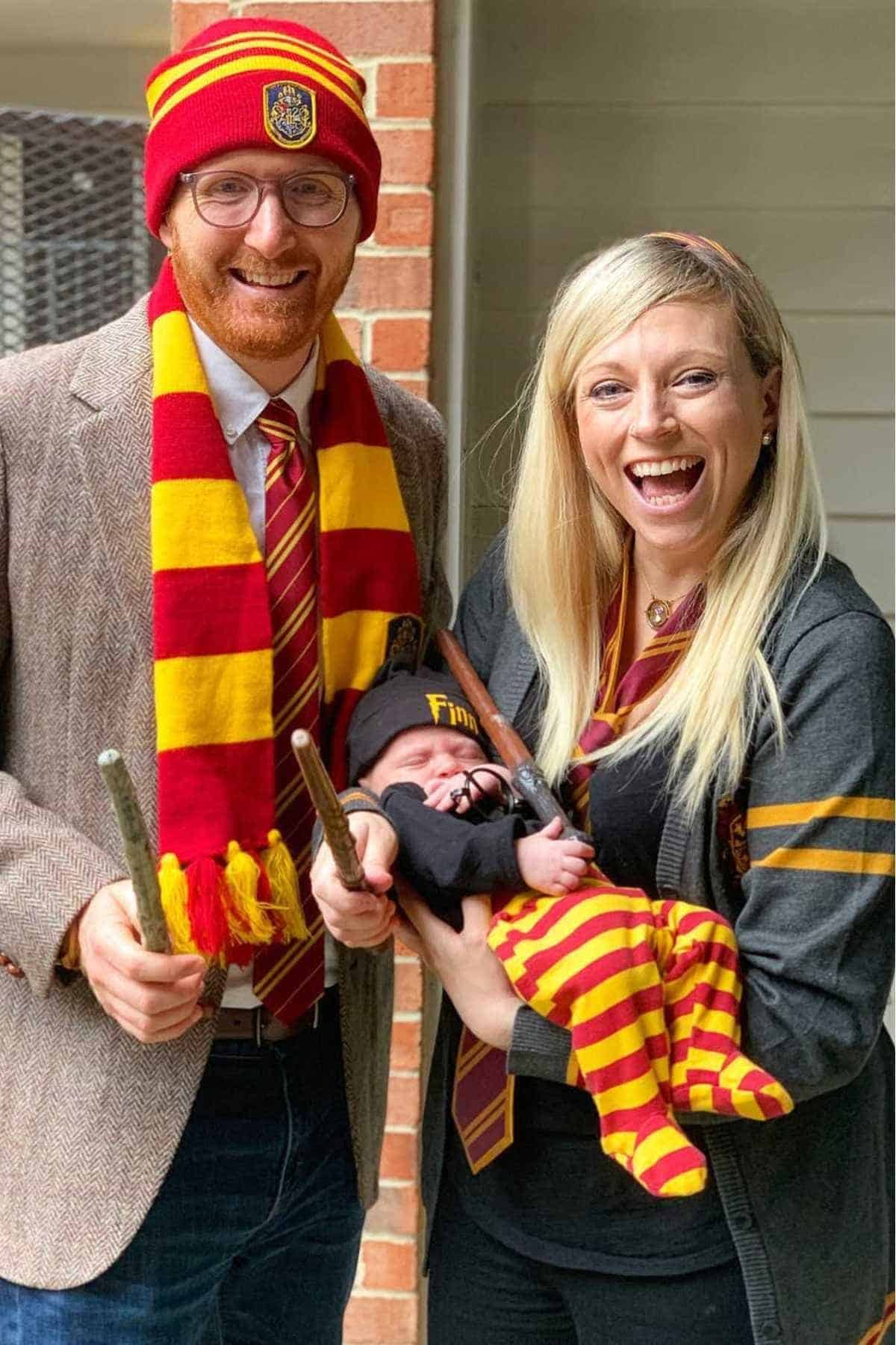 Three people dressed in Gryffindor-themed Harry Potter costumes, with the adults holding wands and the baby wearing a striped hat and pants.