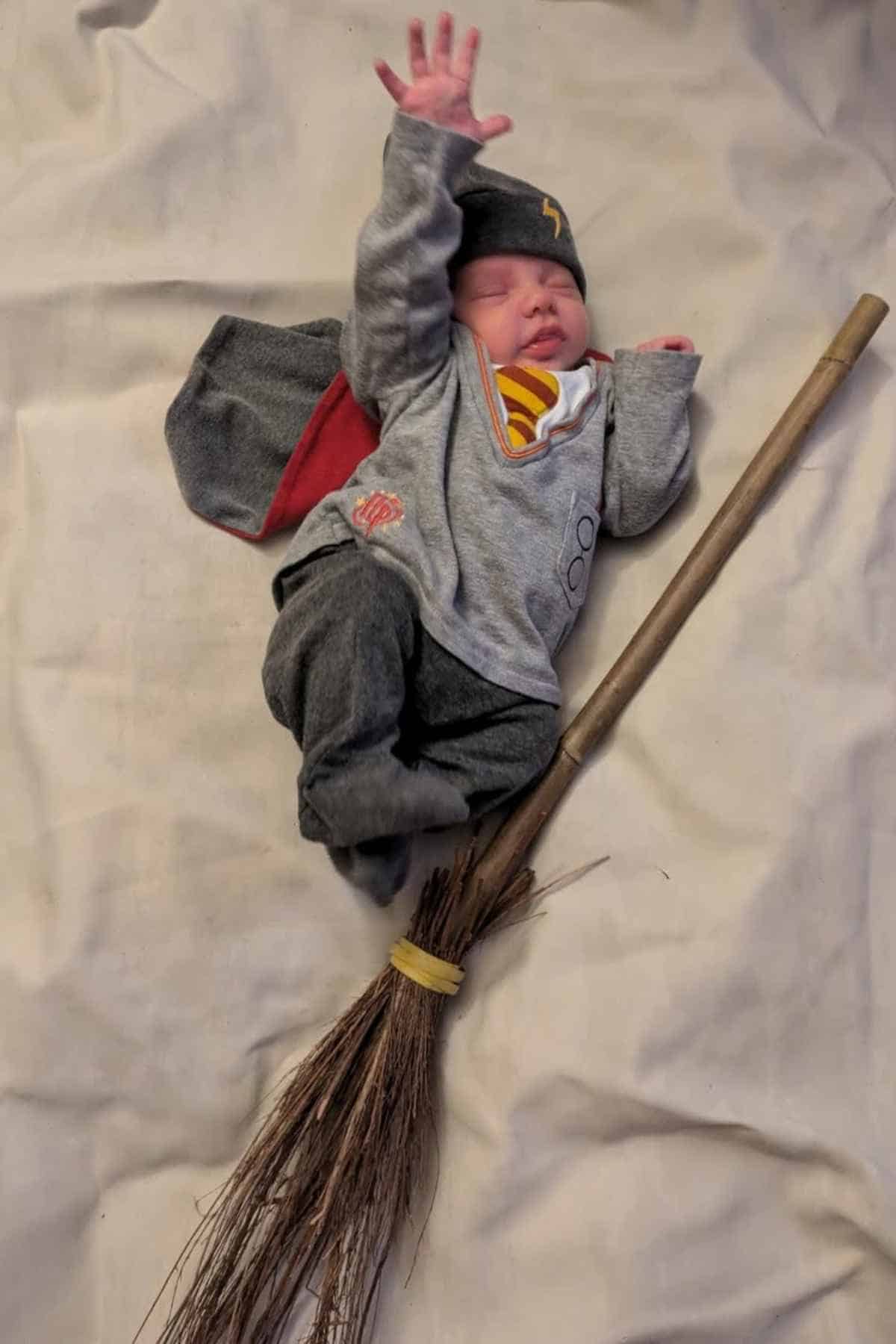 A baby dressed in a superhero outfit lies on a blanket, posed with one arm raised and a broom beneath them, resembling flying on a broomstick.