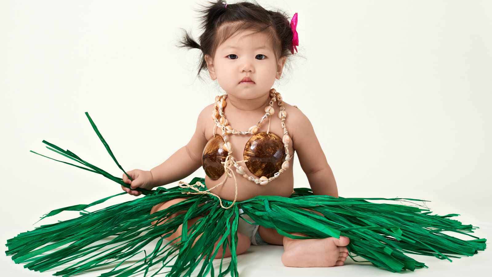 A young child sits on the floor wearing a green grass skirt, shell necklace, and coconut shell top, holding green strands, against a plain white background.