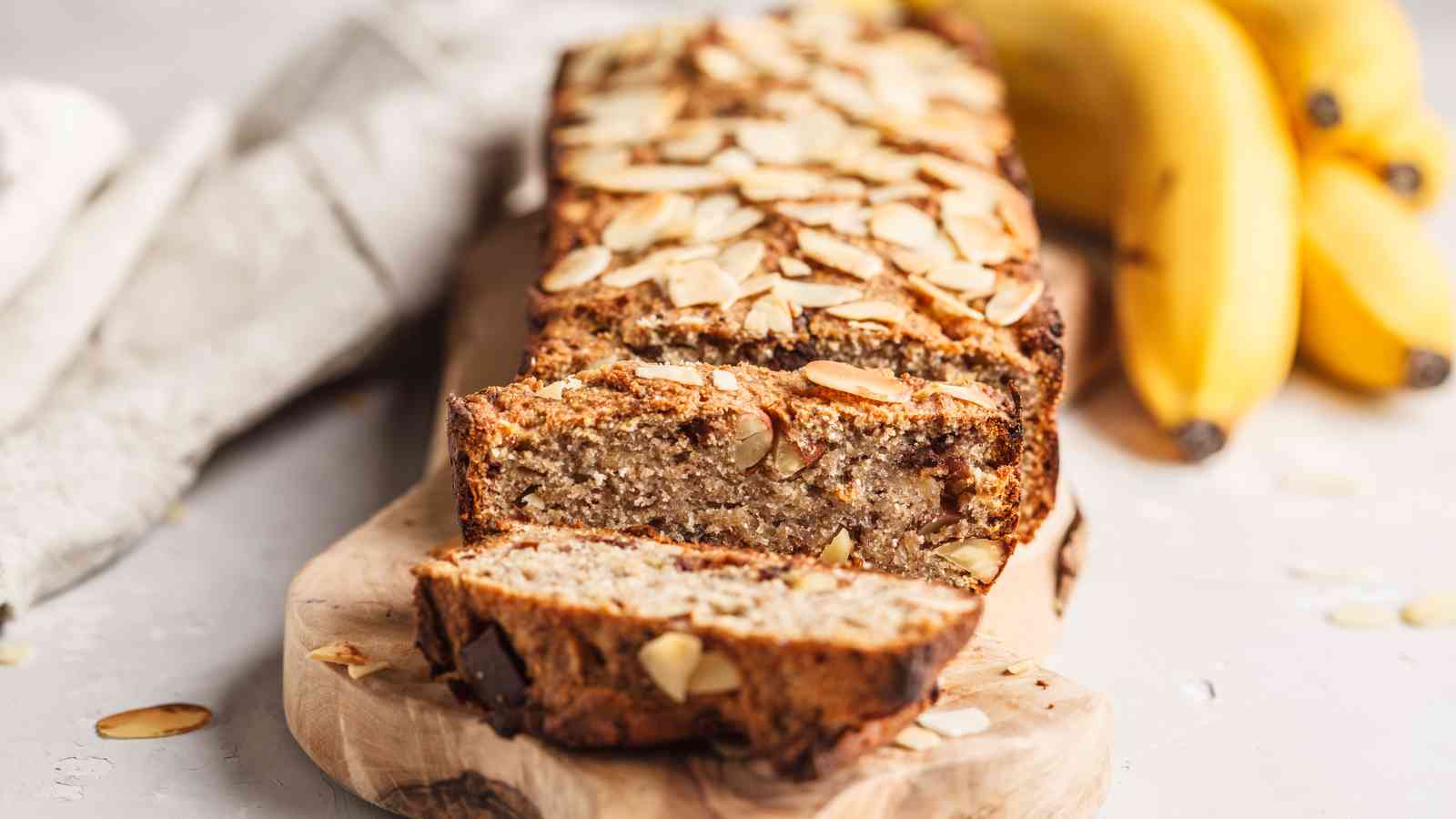 A sliced loaf of banana bread topped with almond slices sits on a wooden board, with bananas in the background.
