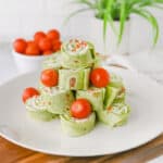 A plate of spinach tortilla pinwheels filled with cream cheese and vegetables, stacked and garnished with whole cherry tomatoes. A bowl of tomatoes and a plant are in the background.