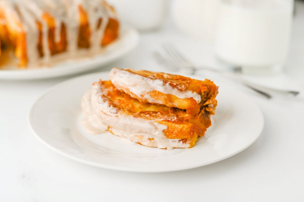 Three slices of pumpkin bread with icing are served on a white plate, with a fork and glass of milk in the background.