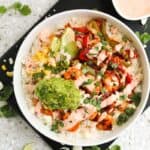 A bowl shrimp fajitas topped with guacamole, corn, sautéed peppers, fresh cilantro, lime wedges, and a drizzle of sauce.