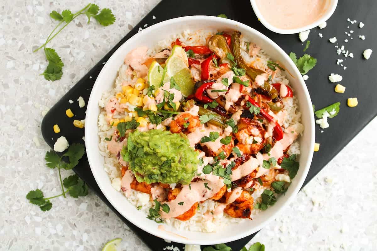 A bowl of rice topped with grilled chicken, guacamole, corn, sautéed peppers, fresh cilantro, lime wedges, and a drizzle of sauce.