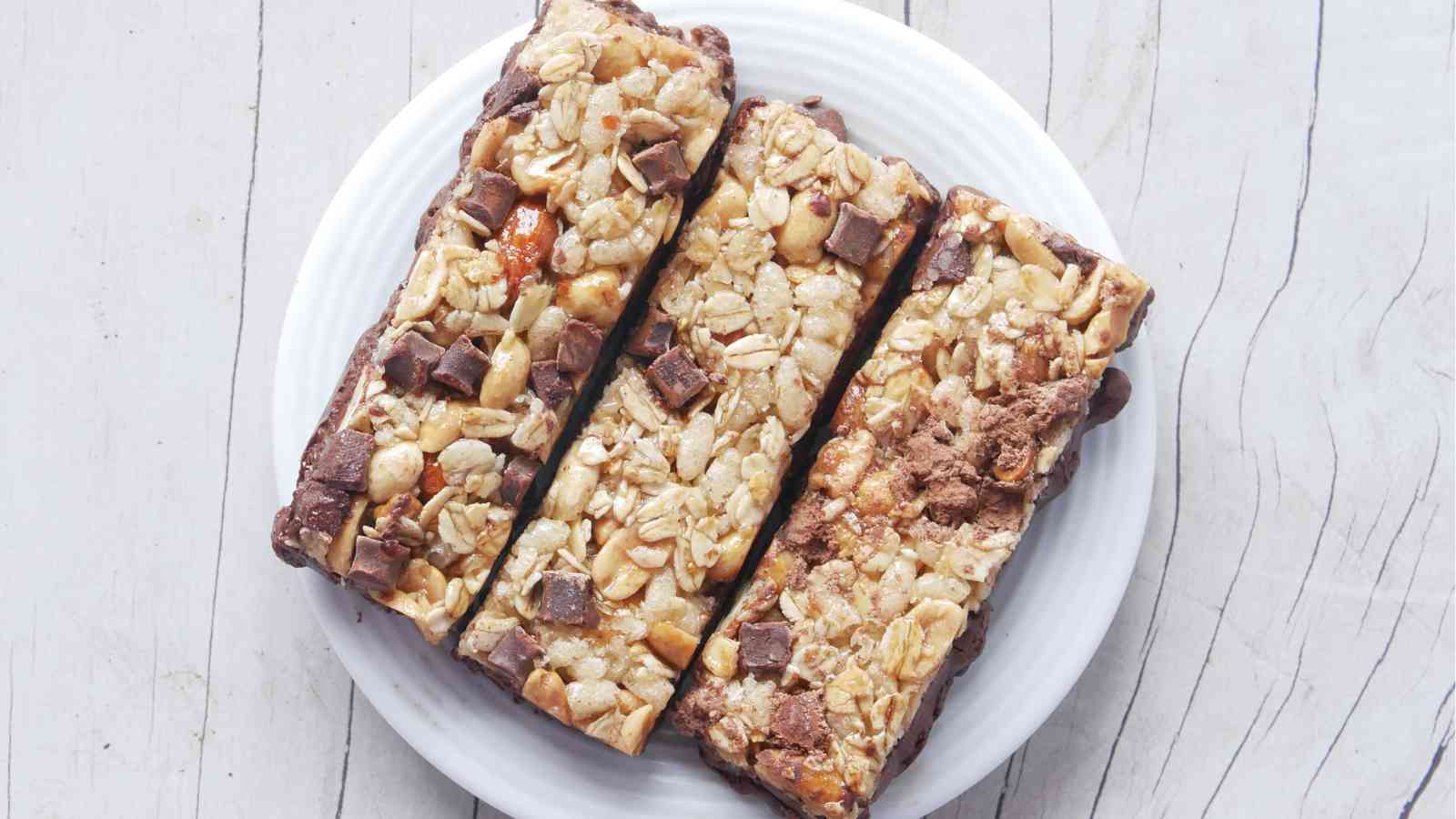 Three granola bars with oats, nuts, and chocolate chunks are arranged side by side on a white plate on a light wooden surface.