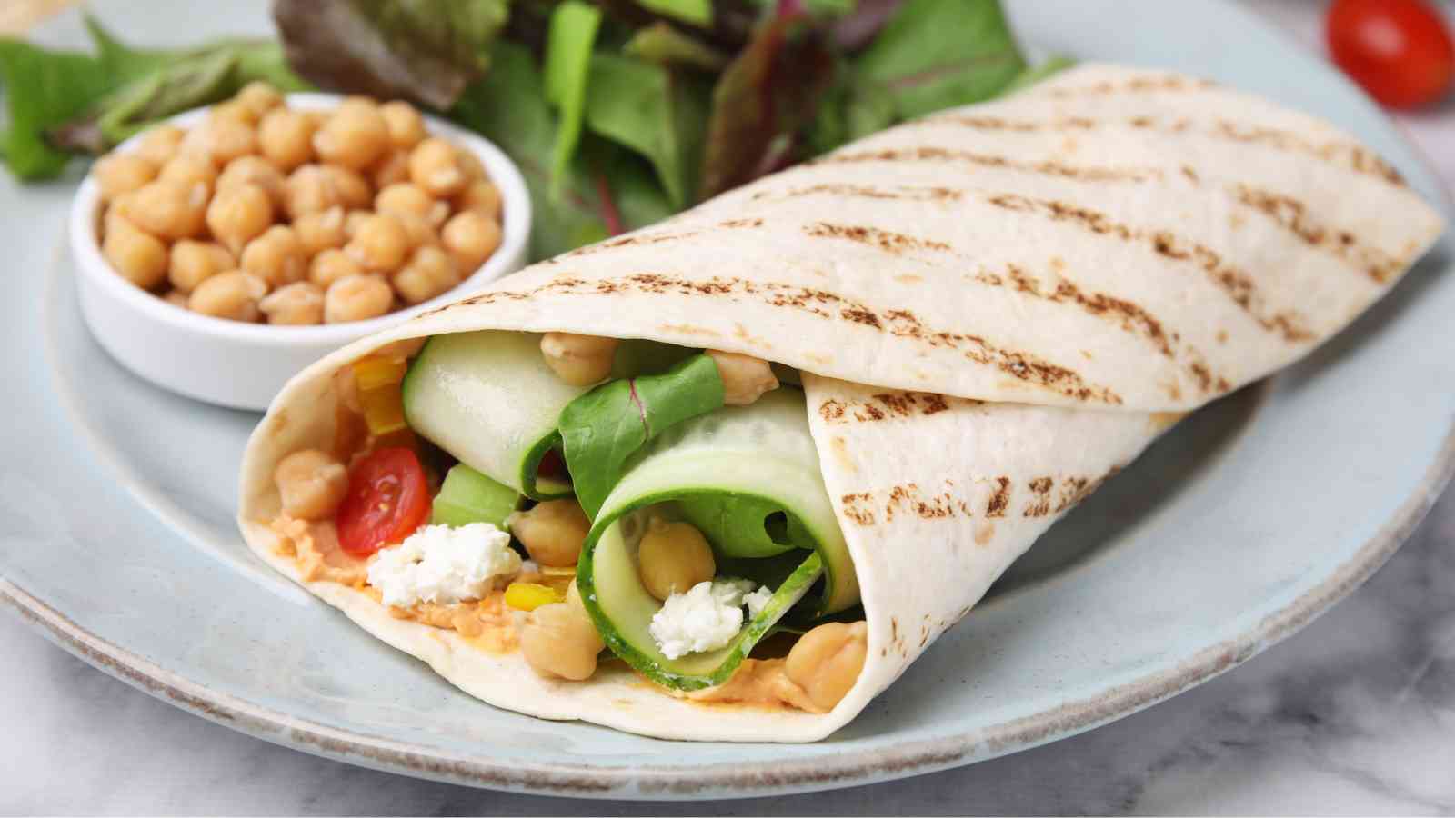 A grilled wrap filled with chickpeas, cucumber, greens, tomatoes, and feta cheese, served with a small bowl of chickpeas and a side salad on a plate.