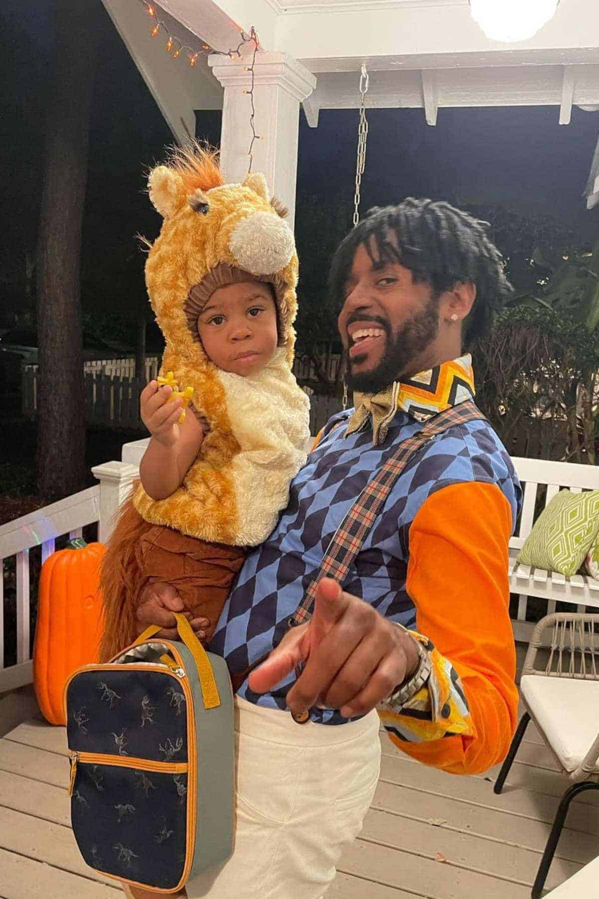 An adult holding a child dressed in a horse costume on a porch; the adult is wearing a colorful shirt and pointing toward the camera.