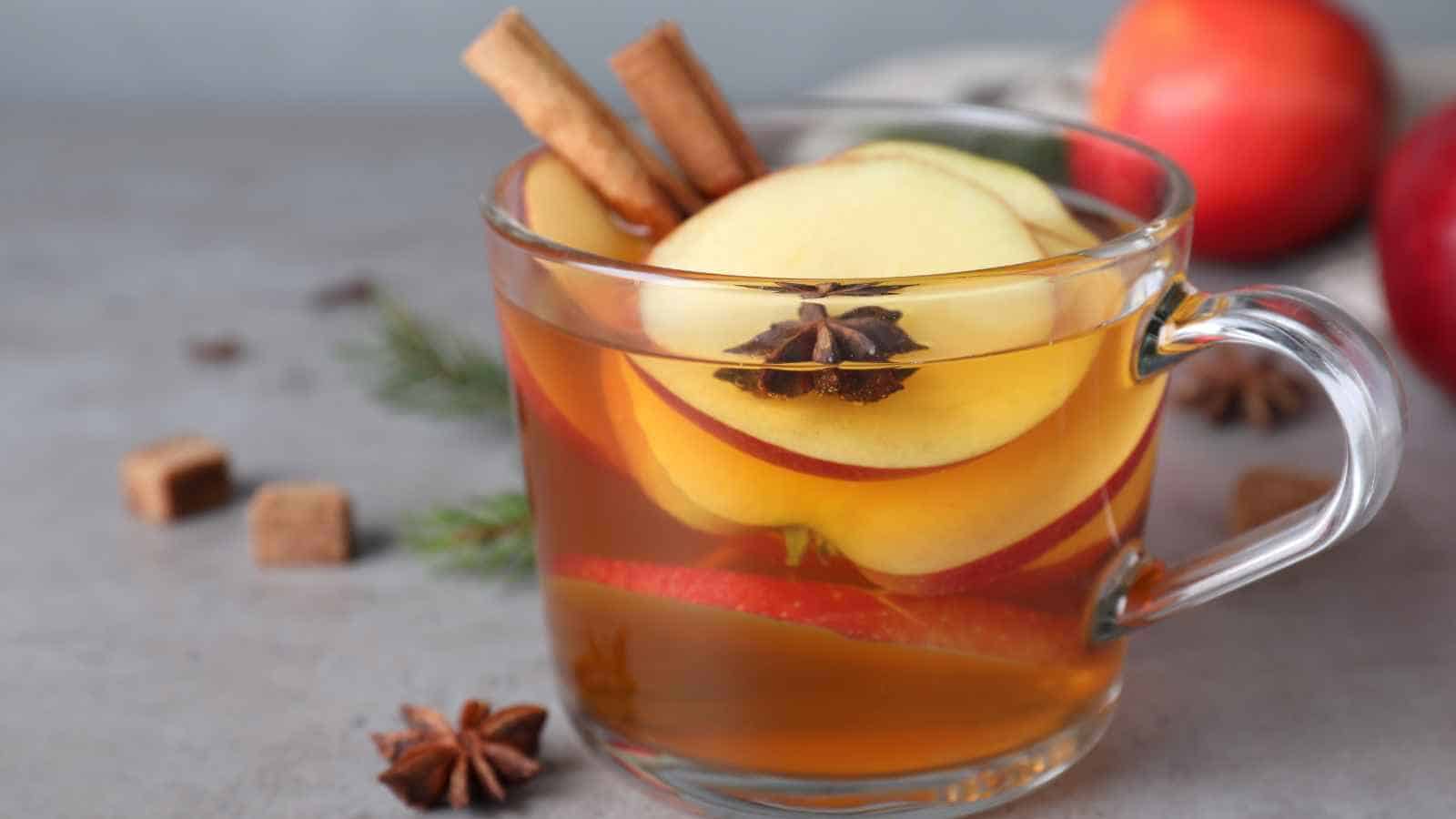 A clear glass mug filled with spiced apple tea, garnished with apple slices, star anise, and cinnamon sticks. Brown sugar cubes and whole spices are nearby.