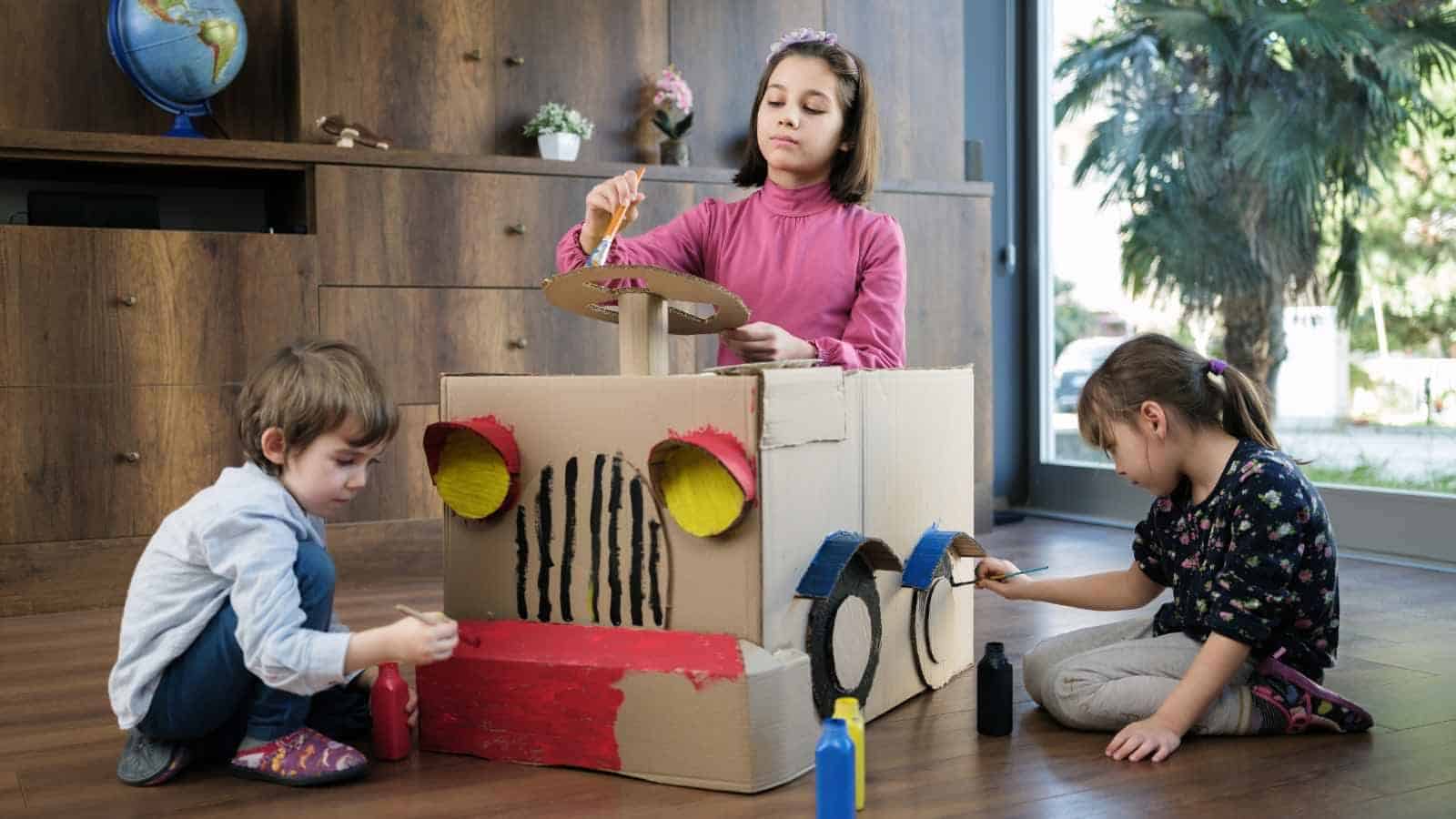 Three children sit on the floor painting and decorating a large cardboard box made to look like a car, using paint and craft materials.