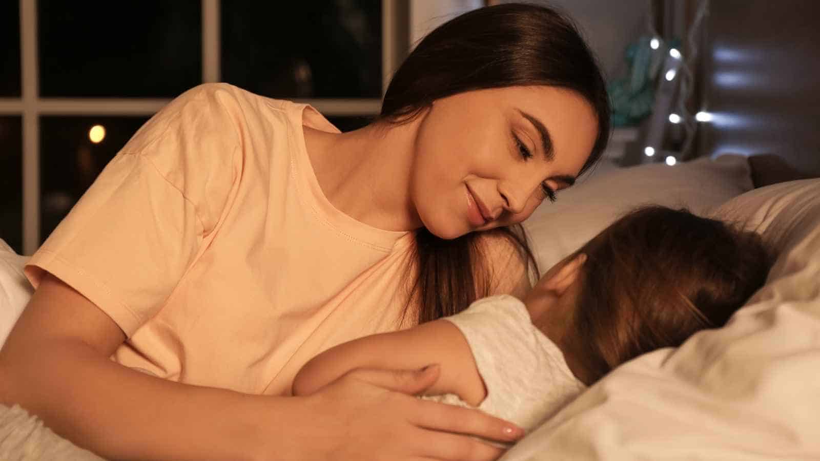 A woman lies in bed, smiling gently at a young child who is resting beside her.