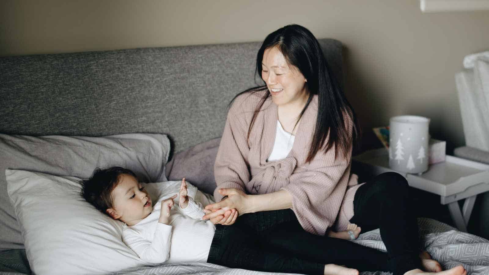An adult and a child are sitting on a bed together; the child is lying down while the adult smiles and holds the child’s hand.