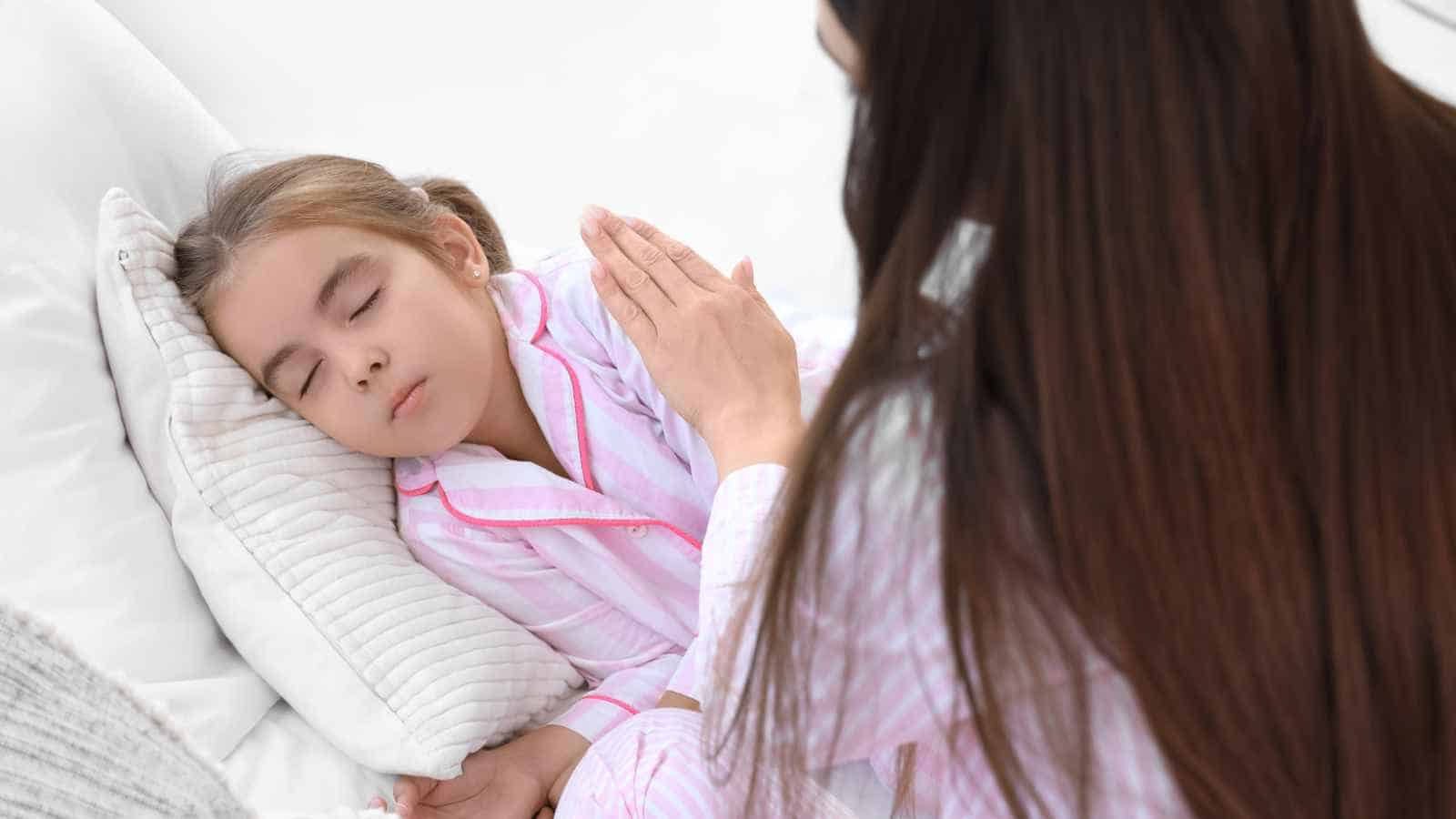 A woman sits beside a sleeping child, placing a hand gently on the child's arm. Both are wearing pink striped pajamas.