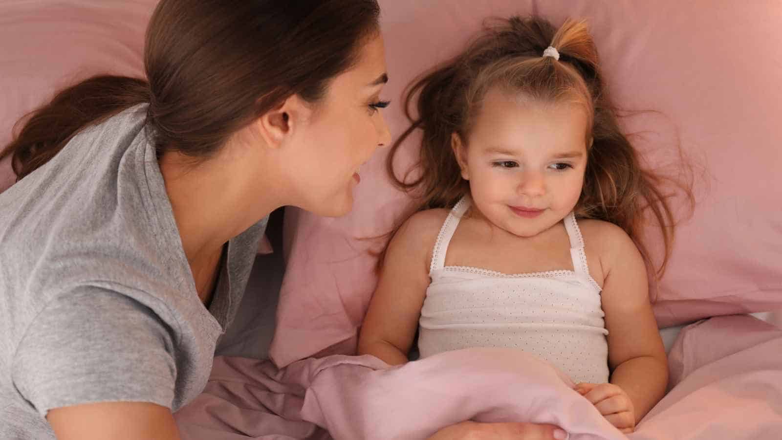 An adult sits beside a young girl lying in bed with pink sheets, both appearing to interact in a calm and gentle manner.