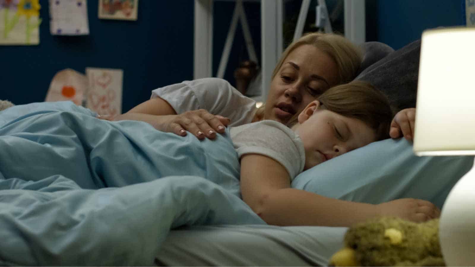 An adult and a child lie in bed under a blue blanket; the adult has a hand on the child's back as both rest, with a lamp and stuffed animal in the foreground.