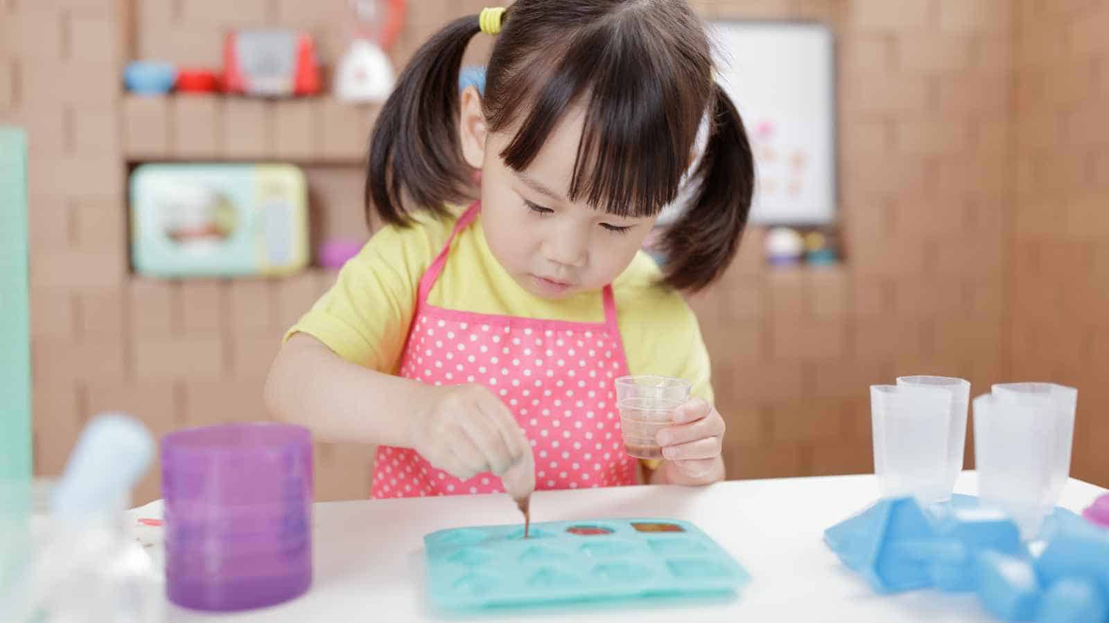Young child in pigtails wearing a pink apron uses a dropper and silicone mold at a table for a science or craft activity.