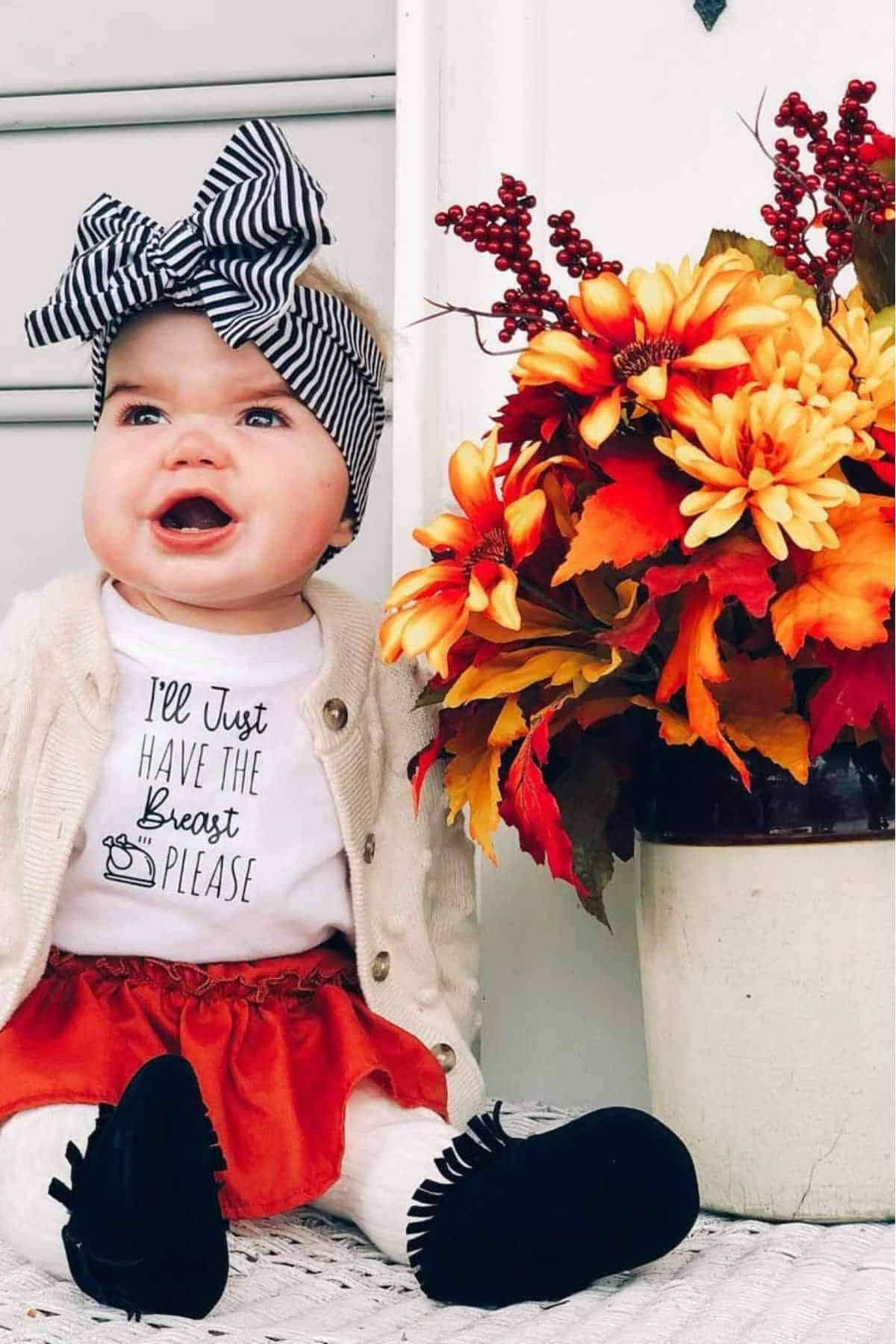 Baby wearing a headband, cardigan, and skirt sits next to a vase of autumn flowers, smiling and looking up.