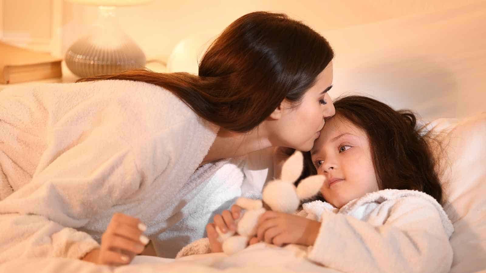 A woman in a white robe kisses a young girl on the forehead as the girl lies in bed holding a stuffed toy.