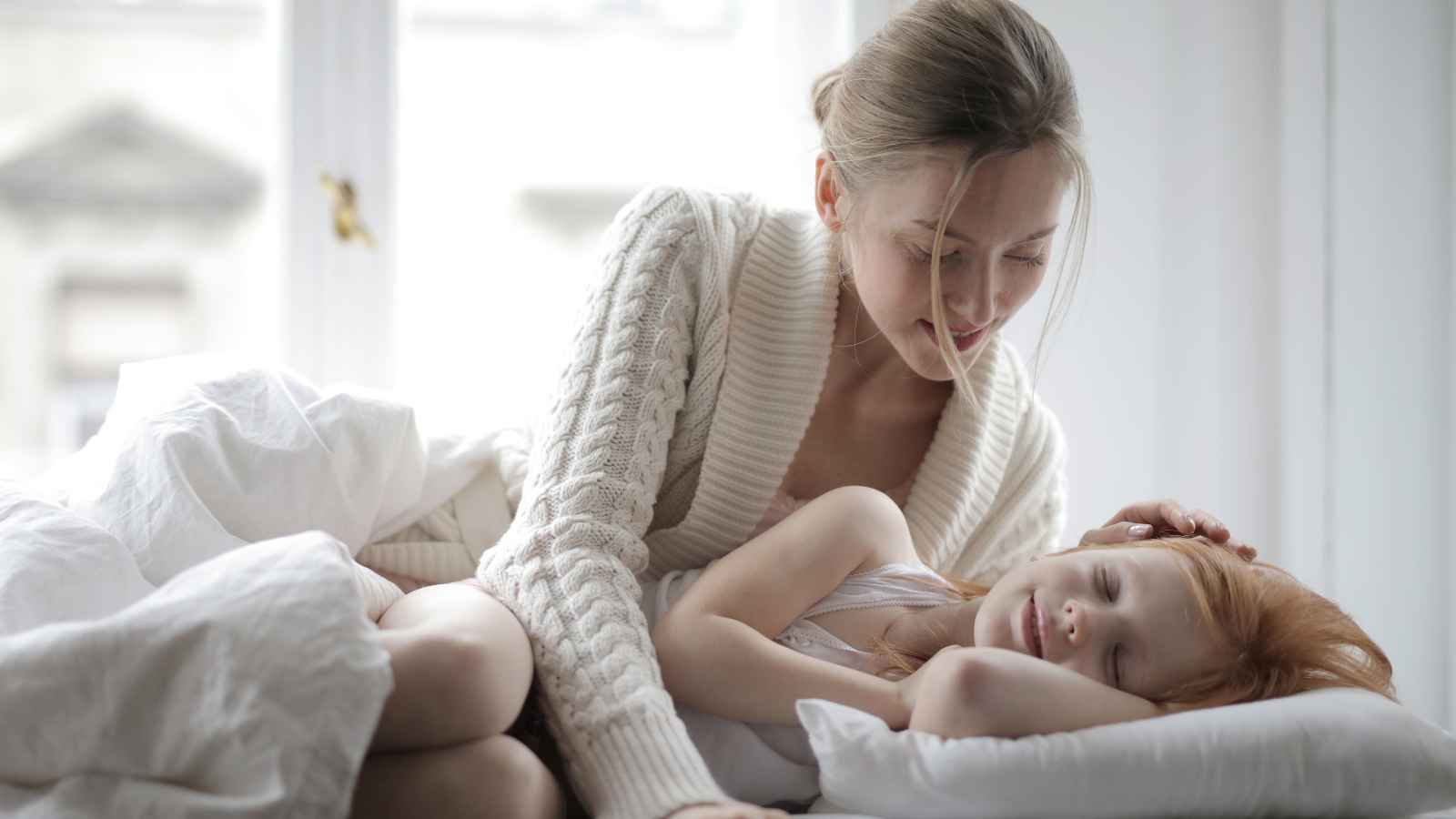 A woman in a white sweater sits on a bed, gently touching the head of a sleeping child with red hair, in a bright, softly lit room.