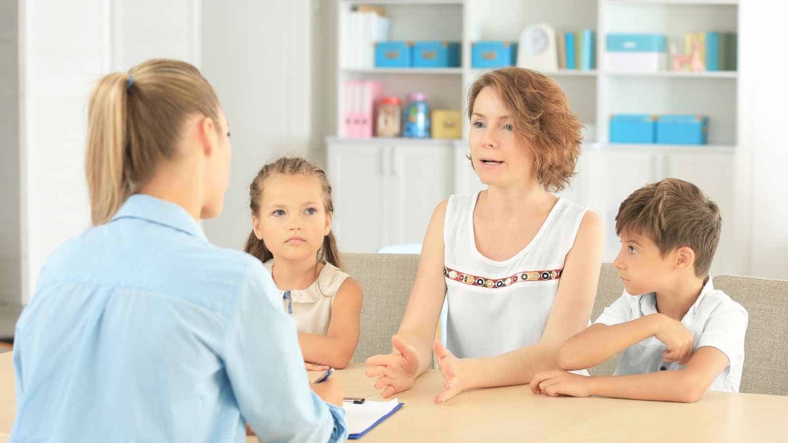 A woman and two children sit across from another woman with a clipboard, having a discussion in a brightly lit office or classroom setting.